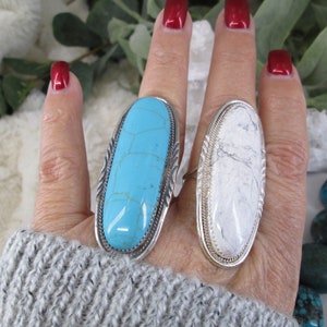 May include: Two silver rings with oval shaped turquoise and white stones. The rings are on a person's finger.