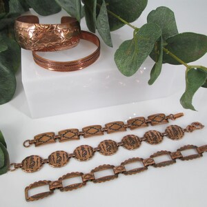 May include: Copper bracelets and a cuff bracelet with etched designs. The cuff has a floral pattern, while the bracelets feature geometric and figure designs. The jewelry is displayed on a white surface with green foliage.
