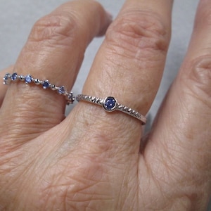 May include: Two silver rings with blue gemstones. The ring on the index finger has a single round gemstone, while the ring on the middle finger has a row of smaller gemstones.