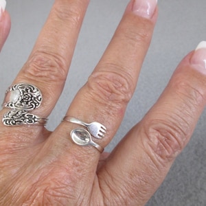 May include: Two silver rings, one with a spoon and fork design and the other with an ornate floral design, worn on a person's finger.