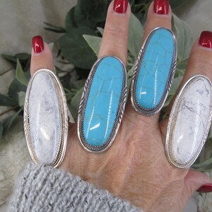 May include: Four silver rings with oval-shaped turquoise and white stones. The rings are worn on a hand with red nail polish.