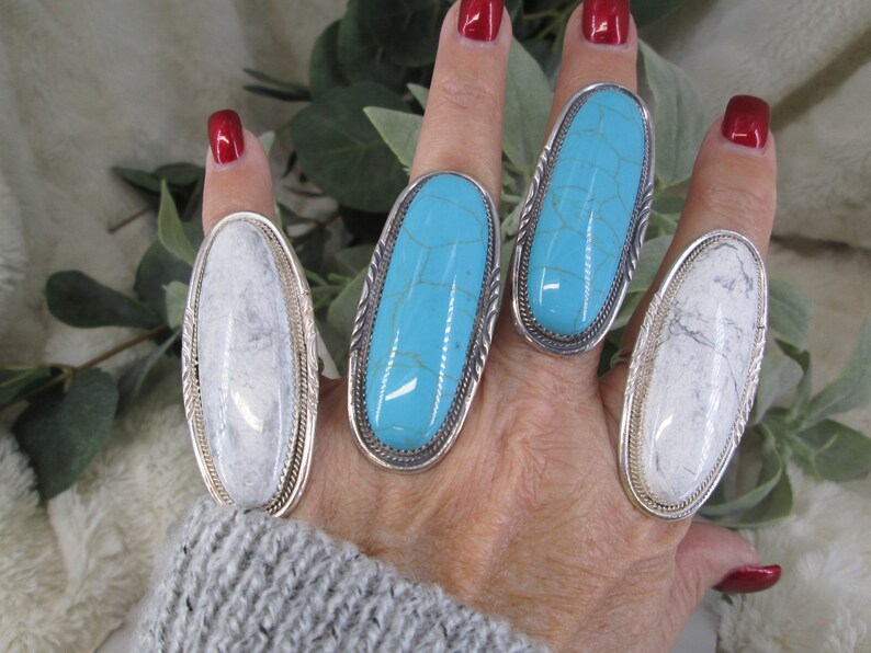 May include: Four silver rings with oval-shaped turquoise and white stones. The rings are worn on a hand with red nail polish.