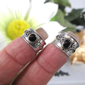 May include: Two silver rings featuring a wide band with a polished finish and decorative scrollwork patterns. Each ring is centered with a small, smooth, oval-shaped black stone set in a twisted metal border, creating a balanced and detailed appearance.