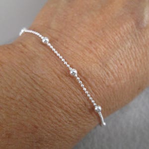 Puede incluir: Una delicada pulsera de plata con una cadena y peque&ntilde;as cuentas redondas espaciadas uniformemente. La pulsera se muestra en una mu&ntilde;eca, destacando su elegancia y dise&ntilde;o sencillos.