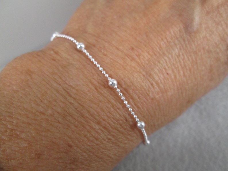 Puede incluir: Una delicada pulsera de plata con una cadena y peque&ntilde;as cuentas redondas espaciadas uniformemente. La pulsera se muestra en una mu&ntilde;eca, destacando su elegancia y dise&ntilde;o sencillos.