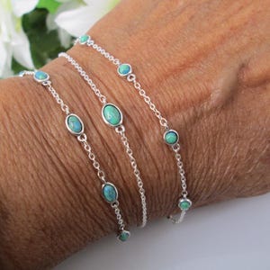 May include: Three delicate silver bracelets with turquoise-colored stones are displayed on a wrist. The bracelets feature oval and round stones, connected by fine silver chains. The wrist is tanned, and a gold watch is visible.