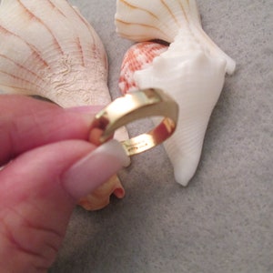 May include: A gold-colored ring with a simple, rounded design. The ring is being held by a hand, with seashells in the background.