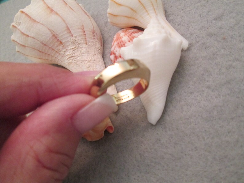 May include: A gold-colored ring with a simple, rounded design. The ring is being held by a hand, with seashells in the background.