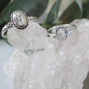 May include: Two silver rings with oval-shaped white stones set in a silver bezel. The rings are on a bed of white crystal rocks.