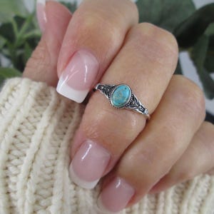 May include: A silver ring featuring an oval turquoise gemstone. The ring has an intricate design around the stone and on the band. The ring is worn on a finger with a French manicure, set against a cream-coloured knitted sweater.
