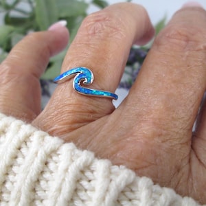May include: A silver ring with a wave design, featuring a blue opal inlay. The ring is worn on a finger, with a white sweater in the background. The wave design is a prominent feature.