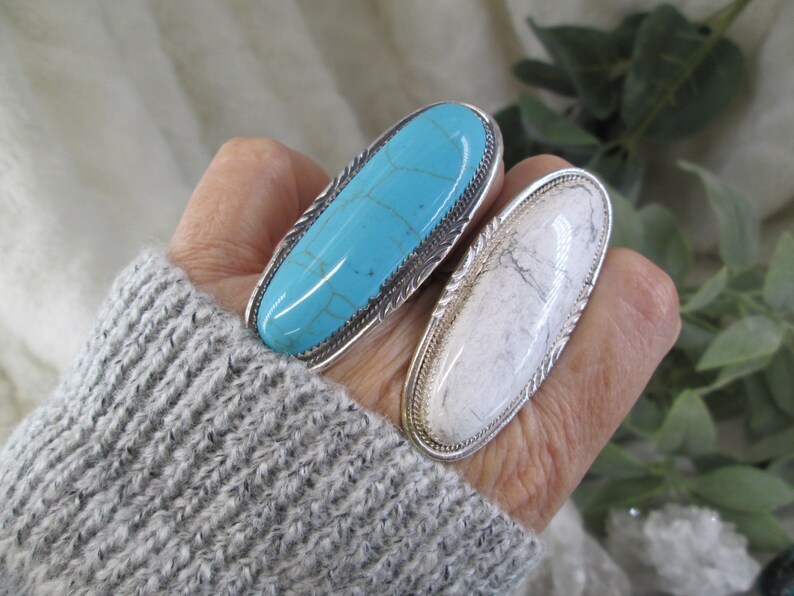 May include: Two silver rings with oval-shaped turquoise and white stones. The rings are worn on a person's fingers.