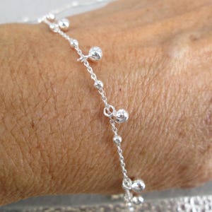 Puede incluir: Un delicado brazalete de plata con una cadena con peque&ntilde;as cuentas redondas y dijes colgantes m&aacute;s grandes. El brazalete se muestra en la mu&ntilde;eca de una persona, destacando su intrincado dise&ntilde;o y sutil elegancia. La joyer&iacute;a es un accesorio simple pero elegante.