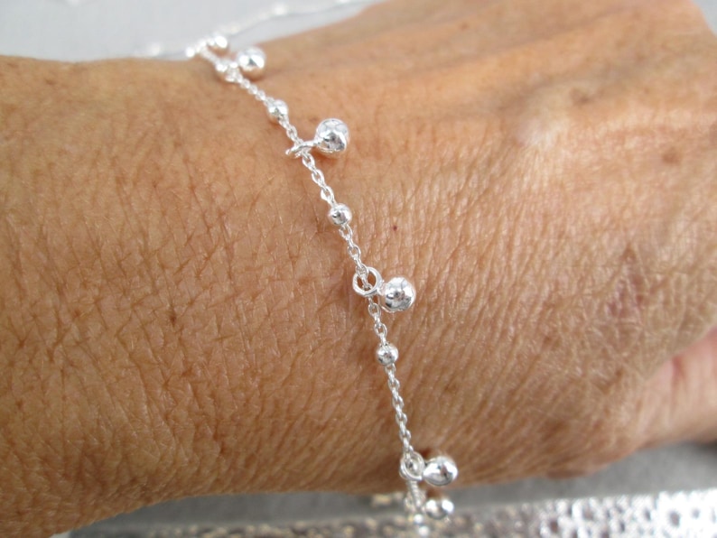 Puede incluir: Un delicado brazalete de plata con una cadena con peque&ntilde;as cuentas redondas y dijes colgantes m&aacute;s grandes. El brazalete se muestra en la mu&ntilde;eca de una persona, destacando su intrincado dise&ntilde;o y sutil elegancia. La joyer&iacute;a es un accesorio simple pero elegante.