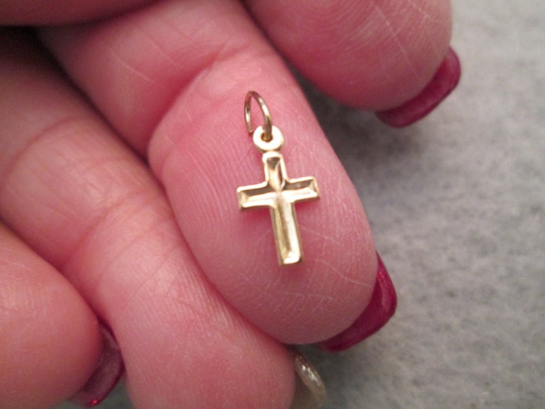 May include: A small gold cross charm pendant. The cross is detailed with a diamond-shaped pattern.