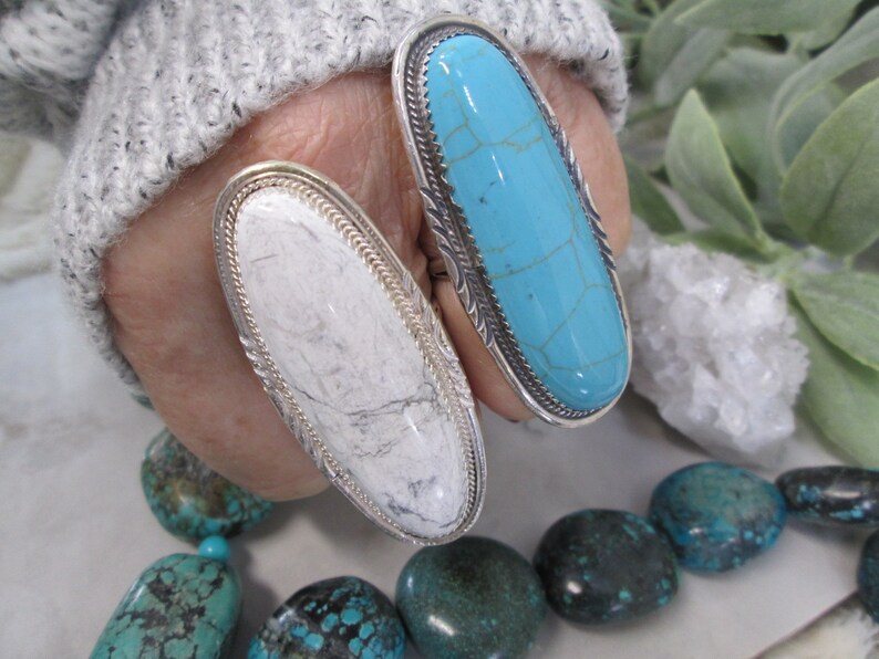 May include: Two silver rings with oval-shaped turquoise stones. The ring on the left has a white stone, while the ring on the right has a blue stone.
