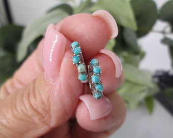 May include: A pair of silver hoop earrings, each adorned with five small, round turquoise stones. The earrings are held up against a blurred green background, showcasing the vibrant blue-green color of the stones.