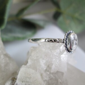 May include: A silver ring with a clear gemstone set in a rope-like bezel. The band is textured with small beads.
