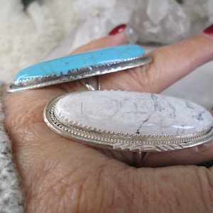 May include: Two silver rings with oval-shaped stones. The top ring has a turquoise stone with a blue and green color. The bottom ring has a white stone with gray veins.