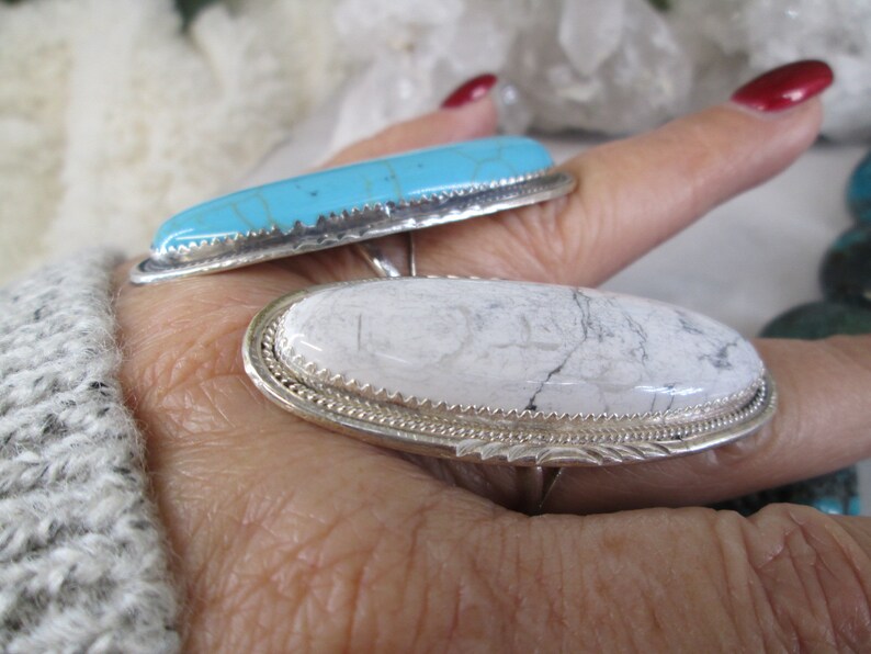 May include: Two silver rings with oval-shaped stones. The top ring has a turquoise stone with a blue and green color. The bottom ring has a white stone with gray veins.