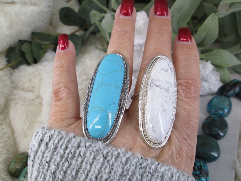May include: Two silver rings with oval shaped turquoise and white stones. The rings are on a person's finger.