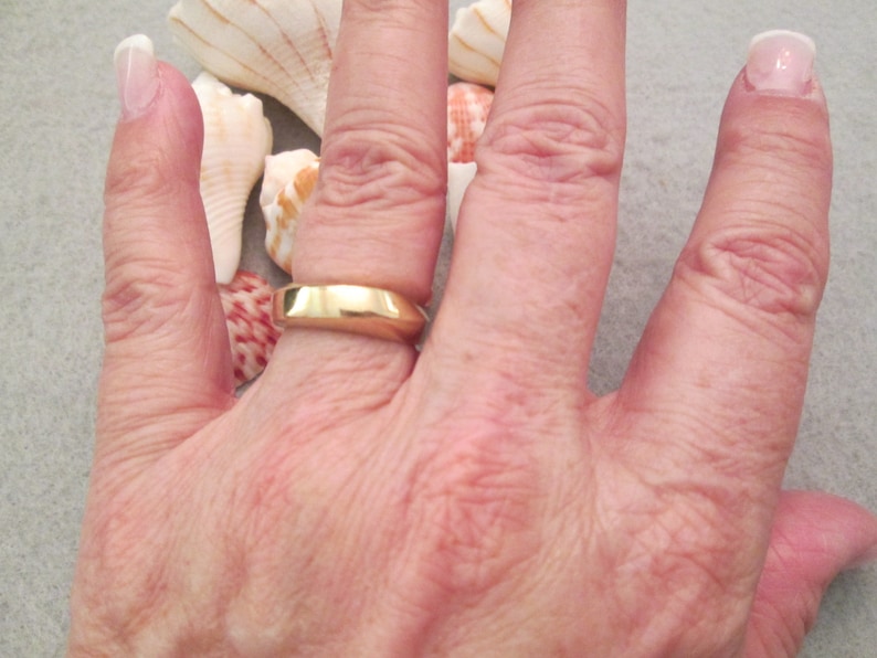 May include: A close-up of a person's hand wearing a gold wedding band. The ring is simple and has a smooth, polished finish.
