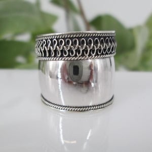 May include: A silver ring with a wide band and a decorative design around the top edge.