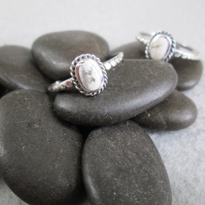 May include: Two silver rings with white oval stones set in a beaded bezel. The rings are displayed on a pile of smooth, grey stones.