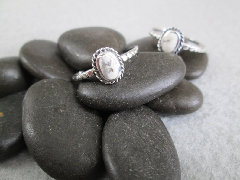 May include: Two silver rings with white oval stones set in a beaded bezel. The rings are displayed on a pile of smooth, grey stones.