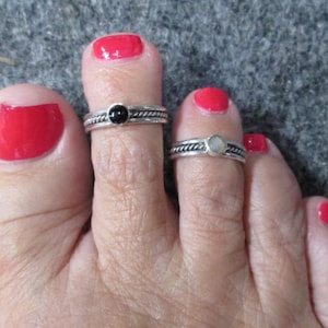 May include: Close-up of a foot with red nail polish and two toe rings. One ring has a black stone, the other a white stone. Both rings have a twisted rope design around the band.