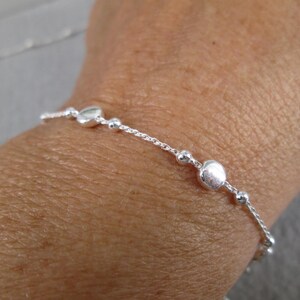 Puede incluir: Una delicada pulsera de plata con una serie de peque&ntilde;as cuentas redondas y cuentas m&aacute;s grandes, aplanadas, en forma de coraz&oacute;n. La pulsera se lleva en la mu&ntilde;eca, mostrando su dise&ntilde;o simple pero elegante. La longitud de la pulsera es de unos 18 cm.
