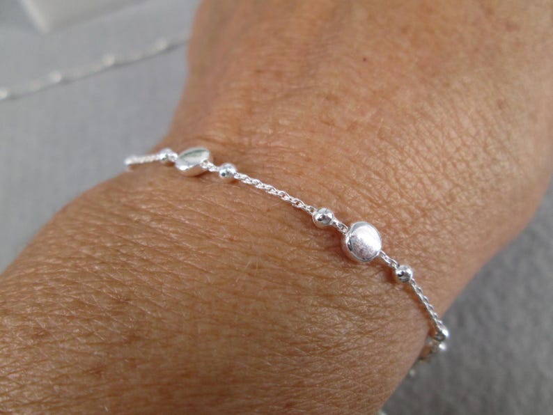 Puede incluir: Una delicada pulsera de plata con una serie de peque&ntilde;as cuentas redondas y cuentas m&aacute;s grandes, aplanadas, en forma de coraz&oacute;n. La pulsera se lleva en la mu&ntilde;eca, mostrando su dise&ntilde;o simple pero elegante. La longitud de la pulsera es de unos 18 cm.