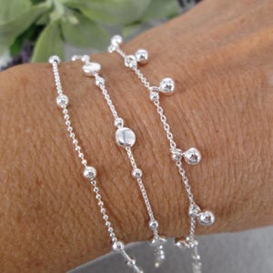 May include: Close-up of multiple silver bracelets with delicate chain links and small, round beads. The bracelets are worn on a person's arm, showcasing the jewelry's design and texture. The bracelets have a simple, elegant design.