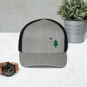 May include: A grey and black trucker hat with a green pine tree and a blue star embroidered on the front.