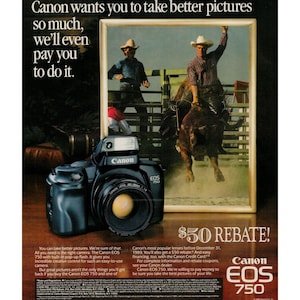 May include: A black Canon EOS 750 camera with a built-in flash. The camera is in focus and the lens is visible. The camera is in front of a framed photo of a cowboy riding a bull. The text "Canon EOS 750" is visible on the camera. The text "$50 Rebate!" is visible in the lower right corner of the image. The text "Canon EOS 750" is visible in the lower right corner of the image.
