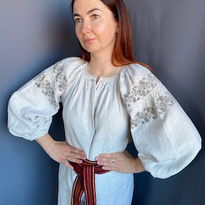 May include: A light beige linen blouse featuring voluminous long sleeves with intricate grey floral embroidery on the shoulders and upper arms. The garment has a gathered round neckline and is cinched at the waist with a woven belt in red, blue, and yellow.