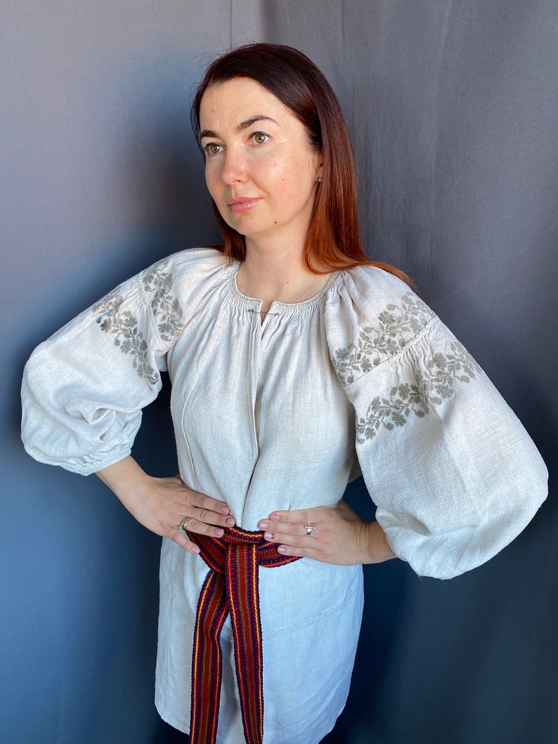 May include: A light beige linen blouse featuring voluminous long sleeves with intricate grey floral embroidery on the shoulders and upper arms. The garment has a gathered round neckline and is cinched at the waist with a woven belt in red, blue, and yellow.