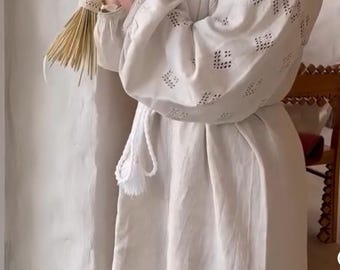 May include: A woman in a white embroidered dress holds a bouquet of dried flowers. She is wearing a pearl necklace and a headband. The background is a white wall with a wooden door.