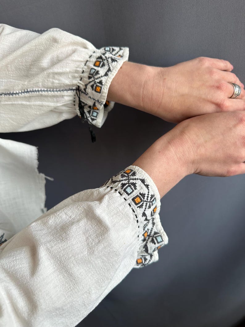 May include: White linen blouse with embroidered cuffs featuring a geometric pattern in black, orange, and blue.