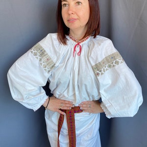 May include: A white embroidered blouse with long sleeves and a red ribbon tied at the neck. The blouse is cinched at the waist with a colorful woven belt.