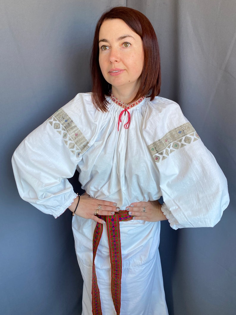 May include: A white embroidered blouse with long sleeves and a red ribbon tied at the neck. The blouse is cinched at the waist with a colorful woven belt.