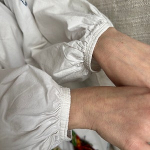 May include: White cotton blouse with long sleeves and a gathered cuff detail. The sleeves are gathered at the wrist and have a simple, elegant design.
