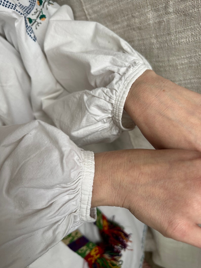 May include: White cotton blouse with long sleeves and a gathered cuff detail. The sleeves are gathered at the wrist and have a simple, elegant design.