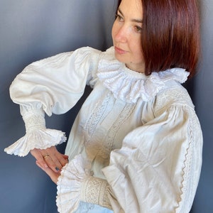 K&ouml;nnte beinhalten: Eine wei&szlig;e Bluse mit einem gro&szlig;en, gekr&auml;uselten Kragen und Spitzendetails an den &Auml;rmeln und am Oberteil.