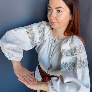 May include: A beige long-sleeved blouse featuring intricate grey floral embroidery along the shoulders and sleeves. The garment has a gathered neckline and elasticated cuffs, paired with a woven belt displaying red, blue, and yellow stripes.