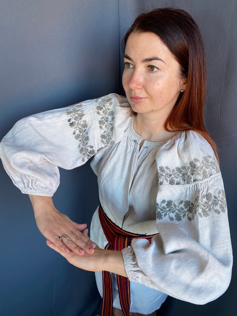 May include: A beige long-sleeved blouse featuring intricate grey floral embroidery along the shoulders and sleeves. The garment has a gathered neckline and elasticated cuffs, paired with a woven belt displaying red, blue, and yellow stripes.