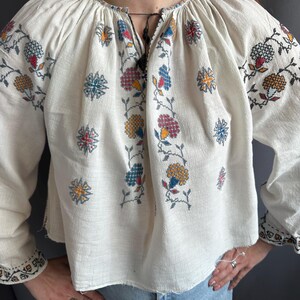 May include: White embroidered blouse with a floral pattern in shades of red, blue, yellow, and green. The blouse has a gathered neckline and long sleeves.