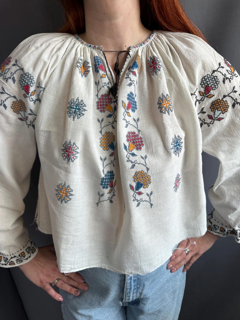 May include: White embroidered blouse with a floral pattern in shades of red, blue, yellow, and green. The blouse has a gathered neckline and long sleeves.