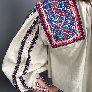 May include: A white linen blouse with colorful embroidered details on the collar, cuffs, and sleeves. The embroidery features geometric patterns in red, blue, green, and black.