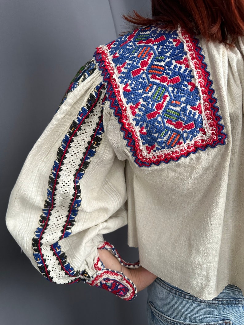 May include: A white linen blouse with colorful embroidered details on the collar, cuffs, and sleeves. The embroidery features geometric patterns in red, blue, green, and black.